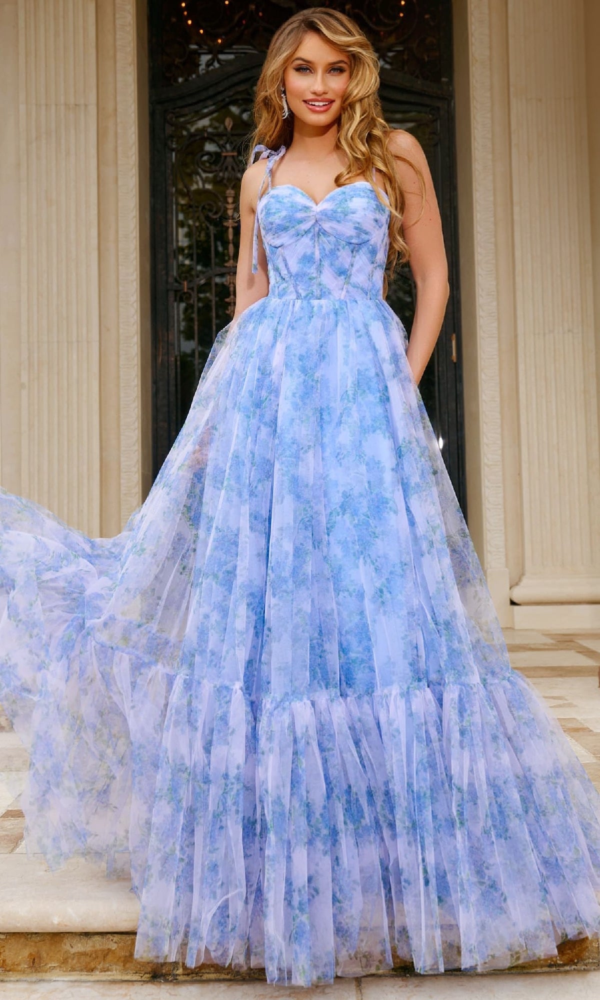 A woman stands before tall columns and a dark ornate door, wearing the Jovani 40432 long prom dress featuring a flowing blue and lavender floral print, sweetheart neckline, and corset bodice.